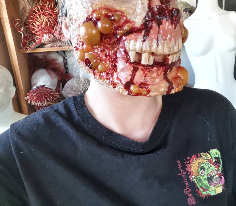 Może przedstawiać: Straszna, realistyczna maska zombie z odsłoniętymi zębami, krwią i krostami. Maska jest noszona na czarnym t-shircie z kolorową grafiką zombie pijącego herbatę i tekstem "#Brainsforte".