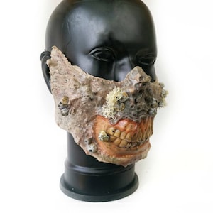 Puede incluir: Una máscara detallada y texturizada con apariencia desgastada. La máscara presenta un diseño de boca abierta en descomposición con dientes visibles y detalles similares a percebes. La paleta de colores incluye tonos marrones, beige y naranja.