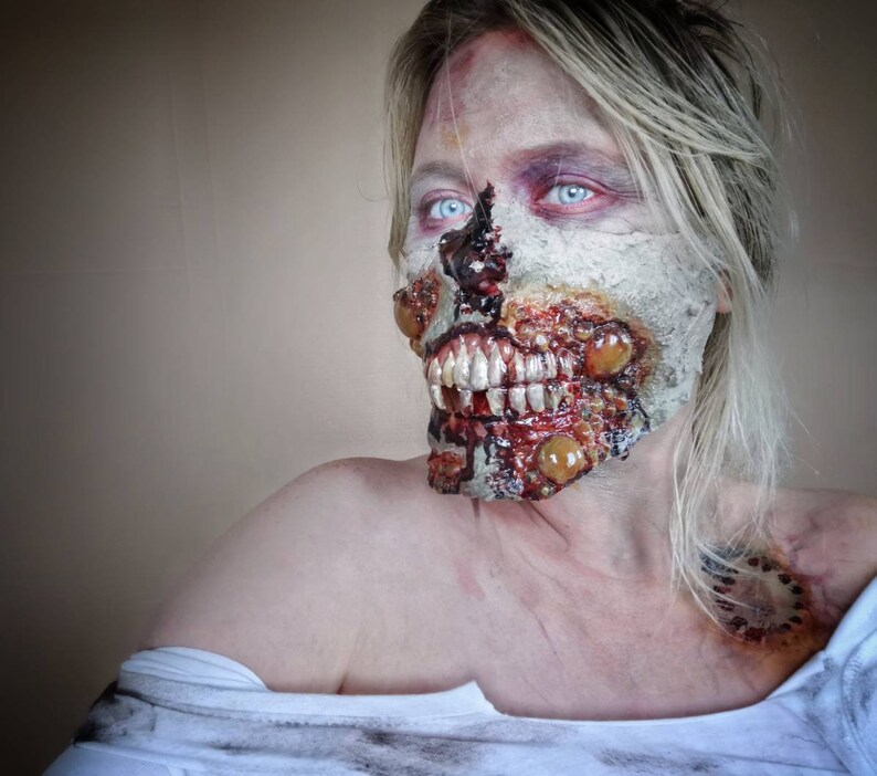 Może przedstawiać: Osoba z makijażem zombie, z odsłoniętymi zębami, ranami i rozkładającym się wyglądem. Makijaż obejmuje bladą cerę, krew i groteskowe detale, tworząc motyw horroru. Osoba ma niebieskie oczy i blond włosy.
