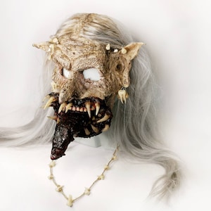 Puede incluir: Una máscara grotesca con un aspecto desgastado y texturizado, con dientes afilados y una boca ensangrentada. La máscara tiene cuencas de ojos blancas, orejas puntiagudas y está adornada con dientes. Un collar de dientes cuelga debajo, sobre un fondo de pelo gris.