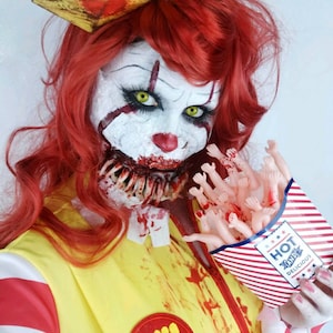 Puede incluir: Un disfraz de payaso aterrador con pelo rojo, pintura facial blanca y un sombrero de barco de papel. El atuendo incluye un chaleco amarillo con rayas rojas y un emblema rojo. El payaso sostiene una caja de extremidades de muñeca.