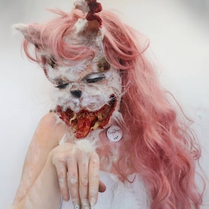 Puede incluir: Una persona con una máscara de gato grotesca con carne expuesta y una etiqueta que dice "Scabby cat". La máscara es blanca y marrón con detalles rojos. La persona tiene el pelo largo y rosa y lleva un atuendo blanco.