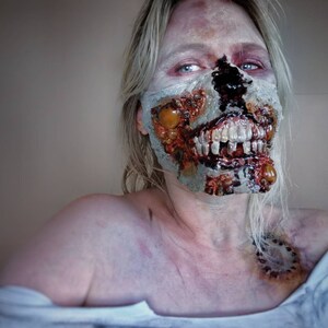 Może przedstawiać: Zbliżenie osoby z realistycznym makijażem zombie. Makijaż obejmuje odsłonięte zęby, rozkładające się ciało i krew. Osoba ma bladą sk&oacute;rę i blond włosy. Całość wygląda strasznie i realistycznie, idealnie na Halloween.
