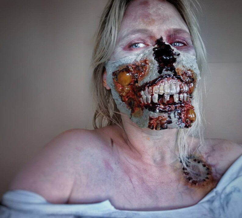 Może przedstawiać: Zbliżenie osoby z realistycznym makijażem zombie. Makijaż obejmuje odsłonięte zęby, rozkładające się ciało i krew. Osoba ma bladą sk&oacute;rę i blond włosy. Całość wygląda strasznie i realistycznie, idealnie na Halloween.