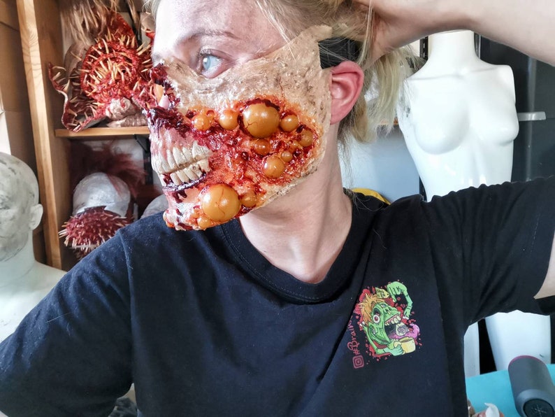 Może przedstawiać: Makabryczna, realistyczna maska zombie z odsłoniętymi zębami, krwawymi ranami i krostami. Osoba nosi czarny t-shirt z kolorową grafiką zombie trzymającego babeczkę. Inne rekwizyty horroru są widoczne w tle.