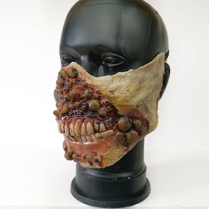 Puede incluir: Una máscara de látex grotesca y realista con dientes expuestos, apariencia de carne en descomposición y numerosas pústulas. La máscara está pintada con tonos marrones, rojos y beige, creando un efecto espantoso. Diseñada para Halloween o eventos de disfraces.