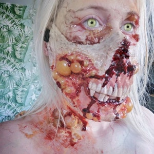 Może przedstawiać: Zbliżenie twarzy z makijażem zombie. Makijaż obejmuje odsłonięte mięso, sztuczne zęby i krew. Osoba ma bladą sk&oacute;rę, zielone oczy i długie, białe włosy. Makijaż jest szczeg&oacute;łowy i realistyczny.