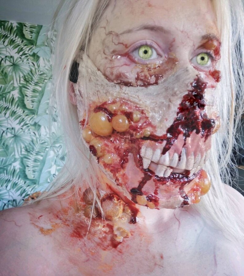 Może przedstawiać: Zbliżenie twarzy z makijażem zombie. Makijaż obejmuje odsłonięte mięso, sztuczne zęby i krew. Osoba ma bladą sk&oacute;rę, zielone oczy i długie, białe włosy. Makijaż jest szczeg&oacute;łowy i realistyczny.