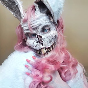 Puede incluir: Una persona con una máscara de conejo grotesca con dientes expuestos y ojos azules. La máscara es blanca y gris, con orejas largas y texturizadas. El pelo rosa y una prenda de piel blanca completan el aspecto inquietante, con uñas rosas.