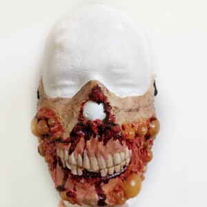 Może przedstawiać: Ręcznie wykonana maska zombie z odsłoniętymi zębami, krwawymi ranami i krostami. Maska jest pomalowana w odcieniach brązu, czerwieni i ż&oacute;łci, tworząc realistyczny i przerażający efekt. Idealna na Halloween lub cosplay.