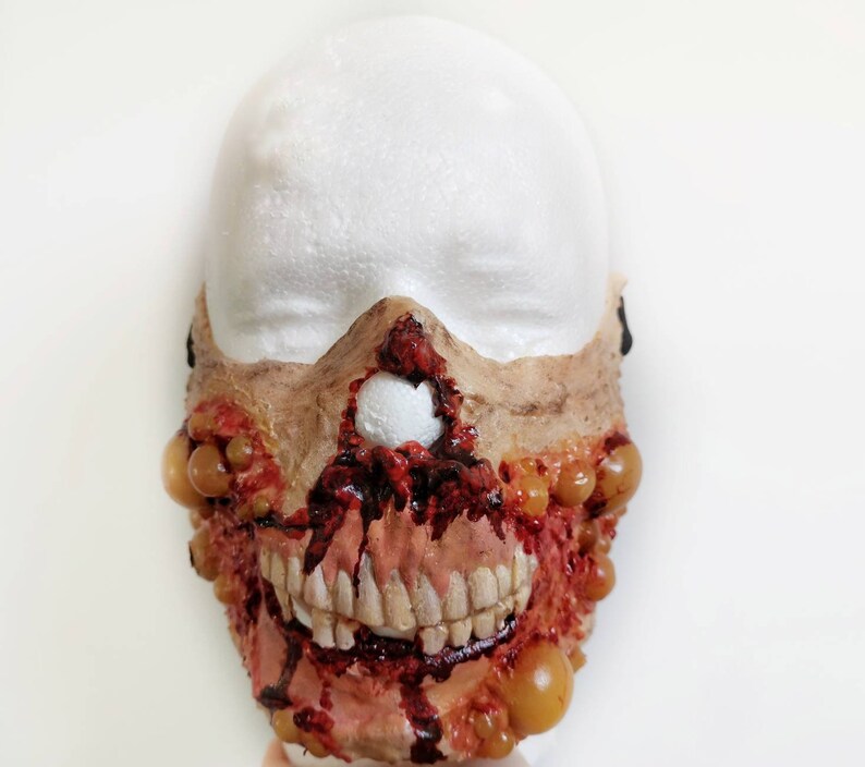 Może przedstawiać: Ręcznie wykonana maska zombie z odsłoniętymi zębami, krwawymi ranami i krostami. Maska jest pomalowana w odcieniach brązu, czerwieni i ż&oacute;łci, tworząc realistyczny i przerażający efekt. Idealna na Halloween lub cosplay.