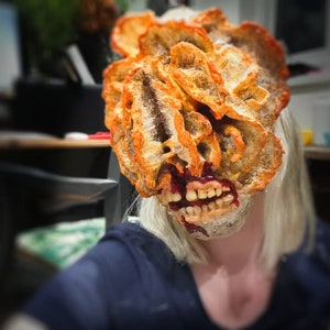 Puede incluir: Una grotesca máscara fúngica naranja y marrón con dientes expuestos y boca ensangrentada. La máscara es un diseño detallado y texturizado, probablemente para un disfraz o cosplay, con un tema zombi o infectado.