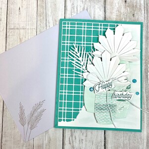May include: A teal green card with a white grid pattern. The card has a white paper cutout of a tropical plant with the words "Happy birthday" on it.