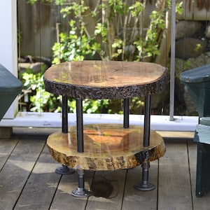 Puede incluir: Una mesa auxiliar de madera de dos niveles, hecha a mano, con borde natural. La mesa presenta una superficie brillante y pulida y está sostenida por tuberías y accesorios de metal negro. La mesa mide aproximadamente 50 cm de diámetro y 46 cm de alto.