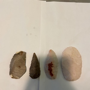 May include: Four stone artifacts arranged in a row. The first is a flat, grey, semi-circular flake. The second is a dark brown, triangular-shaped point. The third is a white, elongated point with a reddish-brown stain. The fourth is a pale, oval-shaped stone.