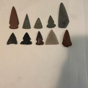 May include: A collection of ten stone arrowheads arranged in two rows. The arrowheads vary in color, including shades of red, gray, black, and brown, and feature distinct triangular shapes with pointed tips and notched or straight bases.