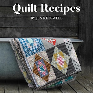 May include: A colorful quilt rests in a metal tub, with the book "Quilt Recipes" by Jen Kingwell in the background. The quilt features various patterned squares in shades of blue, red, gray, and brown. The background is a dark wooden wall.
