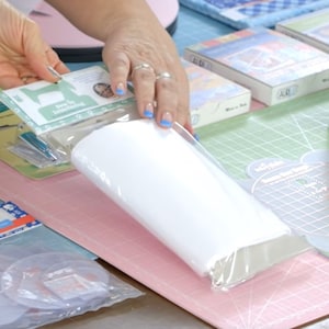 May include: A collection of sewing and quilting supplies, including a roll of white fabric, packaged sewing notions, and a clear plastic ruler with grid lines. The items are arranged on a pink and green cutting mat.