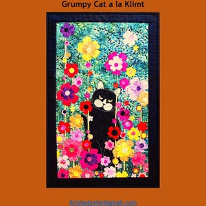 May include: A quilted wall hanging featuring a black cat with an angry expression surrounded by colorful flowers. The artwork is framed in black and set against a teal background. The text "Trouble & Boo Designs" and "Grumpy Cat a la Klimt" are at the top.