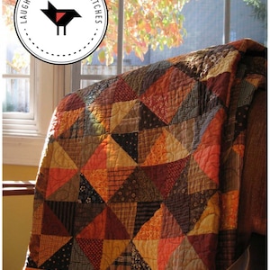 May include: A patchwork quilt in shades of orange, brown, and red, with a geometric pattern of triangles. The quilt is draped over a wooden chair, with a window and fall foliage in the background. The quilt is 52 inches by 64 inches. The text "Laugh Yourself Into Stitches" is on a circular label in the upper left corner of the image.