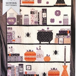 May include: A Halloween-themed quilt with shelves displaying pumpkins, jars, books, and a witch's hat. The quilt features a black cauldron, spiders, and the text "Spooky Shelfie Quilt". The quilt is made with various patterned fabrics.