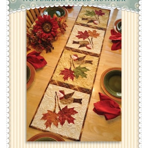 May include: A quilted table runner with a fall theme. The runner features a design of birds and maple leaves in shades of red, orange, yellow, and green. The text "The Vintage Series November Table Runner" is printed on the top of the image.