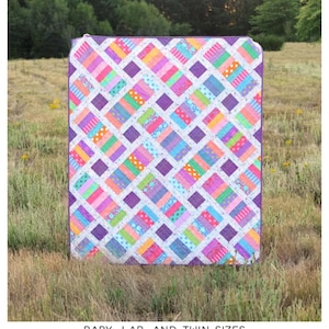 May include: A colorful quilt with a geometric pattern, featuring squares and rectangles in shades of pink, purple, blue, green, and orange. The quilt is displayed outdoors on grass. The text "The Iris Quilt" is at the top.