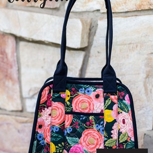 May include: A floral print carryall bag with a dark navy blue trim and handles. The bag features a vibrant pattern of pink and yellow flowers on a dark background. The bottom section is a bright pink material. Text on the bag reads "Boundless Carryall" and "Knot + Thread design".