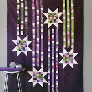 May include: A purple quilt with white star shapes and vertical striped accents in shades of purple, green, and floral patterns. The quilt is displayed on a wall with the text "Showering Stars". A small table with fabric rolls is in the foreground.