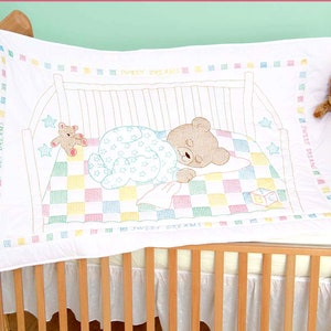 May include: White baby quilt with a brown bear sleeping in a crib. The quilt has a checkered pattern in pink, yellow, green, and blue. The text "Sweet Dreams" is embroidered on the quilt.