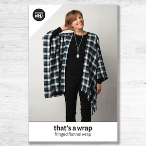 May include: A woman wearing a black top and black pants with a green and white plaid fringed wrap. The wrap has a "that's a wrap" and "fringed flannel wrap" text on the bottom.
