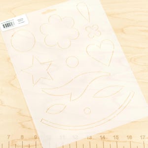 May include: A clear plastic stencil sheet with various shapes cut out, including a flower, star, heart, and circle. The stencil is placed on a light wood surface. The stencil has the text "Laundry Saver Quilt" and "Harmony Stencil".