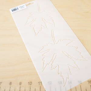 May include: A white stencil featuring two maple leaf designs. The leaves are outlined in a light brown color. The stencil is on a light wood surface. The packaging includes the text "Laundry Basket Quilts" and "Maple Leaves".