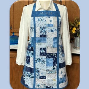 May include: Blue and white floral patchwork apron with the name "JACKIE" embroidered on the bib. The apron is pieced and lined, featuring pockets. The text "Charm Squares Apron" is at the top.