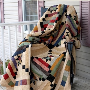 May include: A handmade quilt, named "Roscoe", with a patchwork design. The quilt is 97 x 97 inches and features a variety of colors including beige, red, blue, green, and black. The quilt is draped over a chair on a porch. The website address is visible.