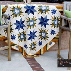 May include: A white quilt with a star pattern in shades of blue, green, and gold is draped over a table. The quilt features large and small stars. Green and striped pillows rest on wooden chairs. The quilt's border has a striped design.
