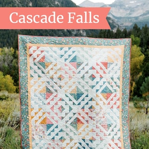 May include: A quilt titled "Cascade Falls" by Amy Smart, Diary of a Quilter, features a geometric pattern of white, yellow, blue, red, and pink triangles and squares against a white background. The quilt is displayed in a field with a mountain range in the background.