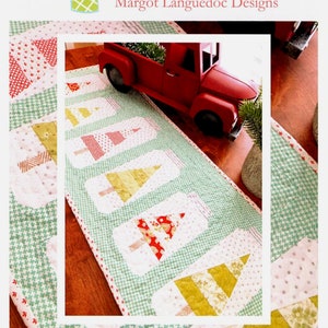 May include: A green and white table runner with a Christmas theme. The runner features six jars with Christmas trees inside. The jars are made of fabric and are sewn together. The runner is called "Christmas Jars" and is a table runner. The website address is www.thepatternbasket.com.