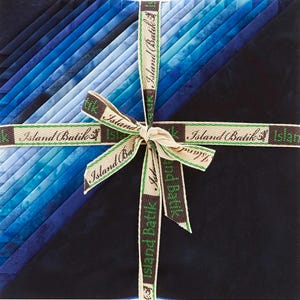 May include: A stack of blue fabric squares, ranging in shades from light to dark, tied with a ribbon. The ribbon is beige with green and brown text that reads "Island Batik". The fabric appears to be cotton and is neatly arranged.