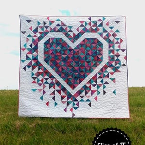 May include: A white quilt with a large, colorful heart design made of patchwork triangles. The heart is surrounded by smaller triangles that appear to be exploding outward. The quilt is displayed on a grassy field with a blue sky in the background. The text "Exploding Heart" is at the top of the image and "Slice of Pi Quilts" is at the bottom.