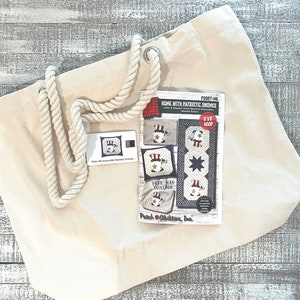 May include: A white canvas tote bag with rope handles. The bag is lying on a wooden surface. A white card with the text "P295T-ME HOME WITH PATRIOTIC GNOMES" is next to the bag. The card shows a design for a patriotic gnome embroidery project.