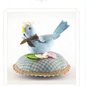 May include: A blue felt bird with a brown bow and white flowers sits on a blue and white checkered pincushion with a lace trim. The pincushion is surrounded by green leaves. The image is titled "Bitty Bird".