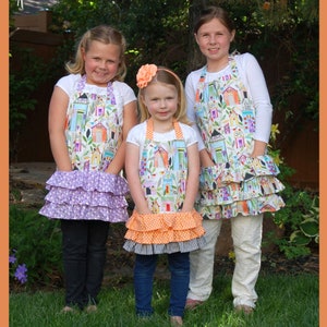 May include: Three girls wearing colorful aprons with a house and bird print. The aprons have multiple ruffles. The girls are standing in front of a green lawn and a wooden fence. The text "Little Lady" and "AbbeyLaneQuilts.com" is at the bottom of the image. The number "#210" is in the bottom right corner.