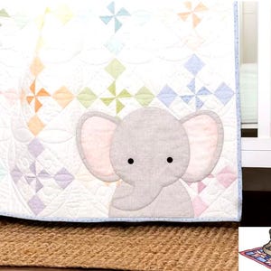 May include: A white baby quilt with a colorful pinwheel pattern and a gray elephant applique. The quilt has a light blue border and is placed on a brown woven rug. A white crib is in the background.
