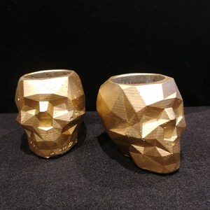 Puede incluir: Dos macetas de calavera geométricas doradas. Las macetas están hechas de hormigón y tienen un acabado mate.