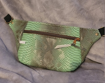 May include: A green snakeskin-patterned fanny pack with a brown zipper and a black strap. The bag is on a gray fuzzy surface.