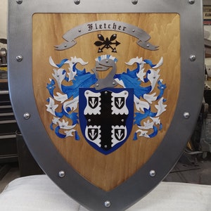 Op de afbeelding: Een houten schild met een metalen frame en klinknagels. Het schild toont een familiewapen met de naam "Fletcher" op een spandoek. Het wapen bevat blauwe, witte en zwarte heraldische elementen, waaronder een helm en gestileerde bloemmotieven.
