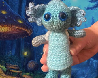 May include: A handmade crochet doll in shades of blue and gray, held in a hand. The doll has large black eyes with blue rims, small arms, and a light blue dress. The background is a whimsical forest scene.