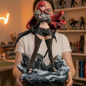 May include: A collectible anime-style figure of a character with long red hair, wearing a dark cropped top, black shorts, and tall boots. The figure stands on a rocky base with a sword at the hip and a flowing red cape. The base features the text POLYGONEER.
