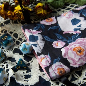 Pode incluir: Um conjunto de dados marmorizados em tons de verde-azulado, branco e dourado, juntamente com uma bolsa de dados floral. A bolsa apresenta um fundo preto com flores de peónia rosa e branca e um design de caveira. Uma vela acesa e flores secas também estão presentes.